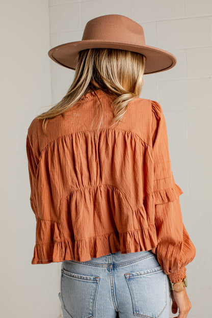 A woman viewed from behind wearing a terracotta orange ruffled blouse, a brown wide-brim hat, and light-wash blue jeans against a white brick wall.