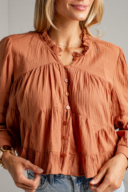 A woman wearing a terracotta rust-colored crinkle gauze blouse with tiered ruffles and a button-down front, paired with blue jeans.