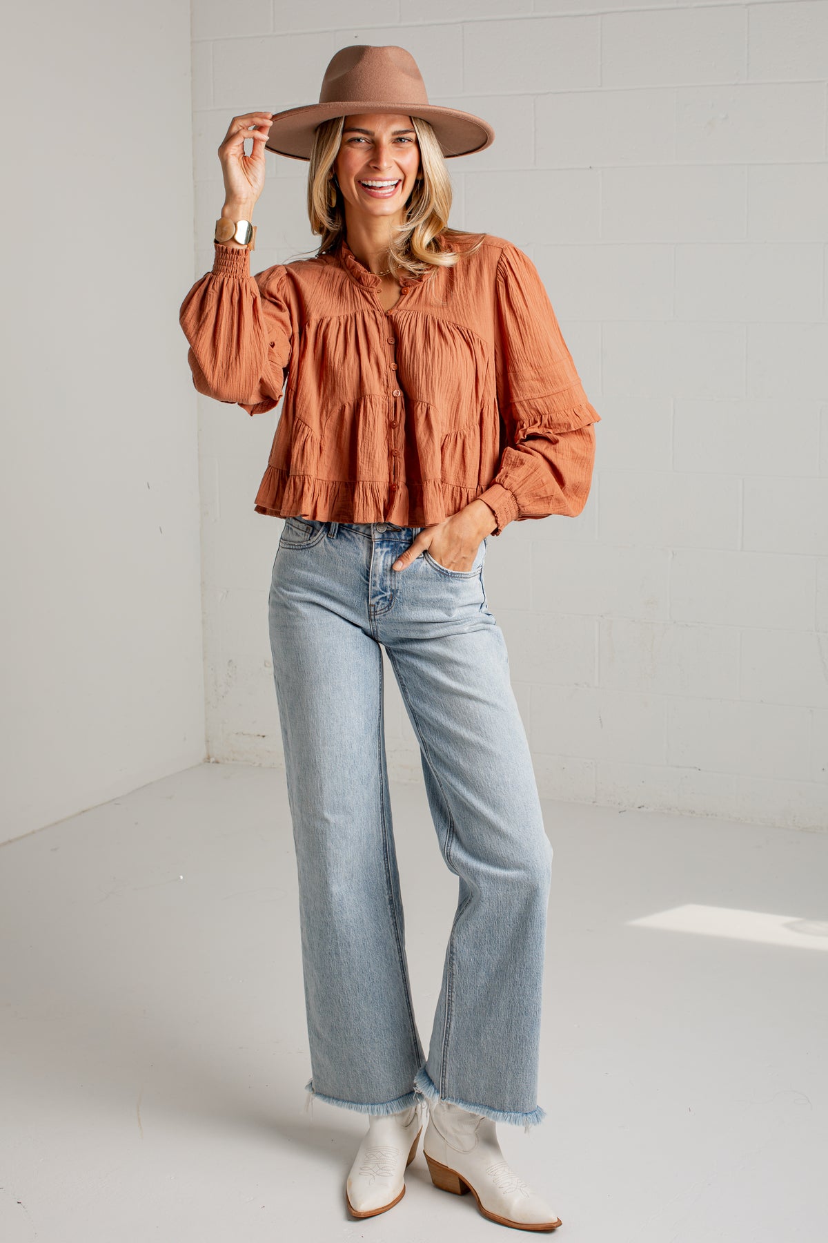 A woman modeling a rust-orange ruffled blouse, light-wash wide-leg jeans, white ankle boots, and a tan wide-brim hat in a bright white studio setting.