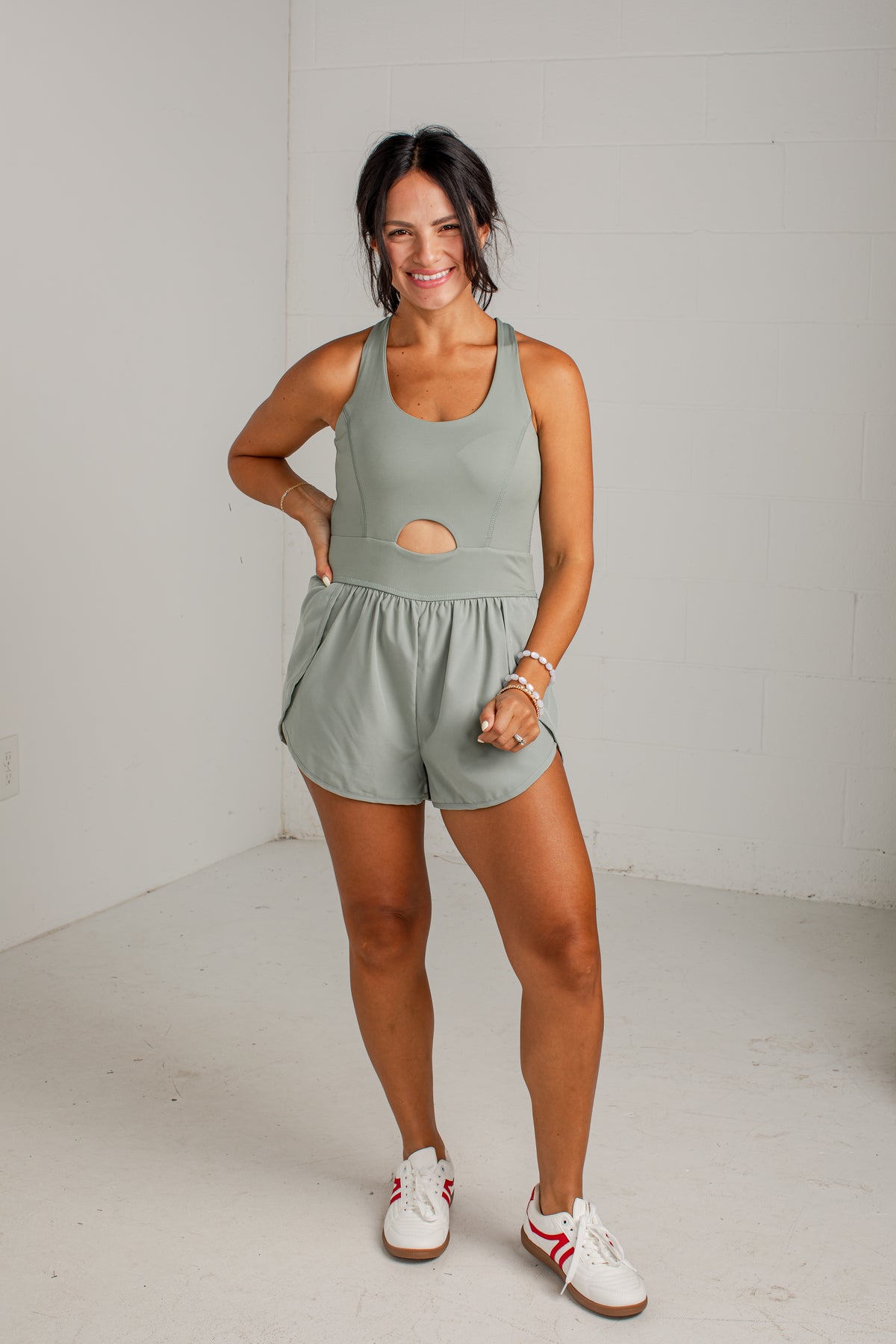 A woman modeling a sage green athletic romper with a cutout at the waist, paired with white sneakers, standing against a white brick wall.