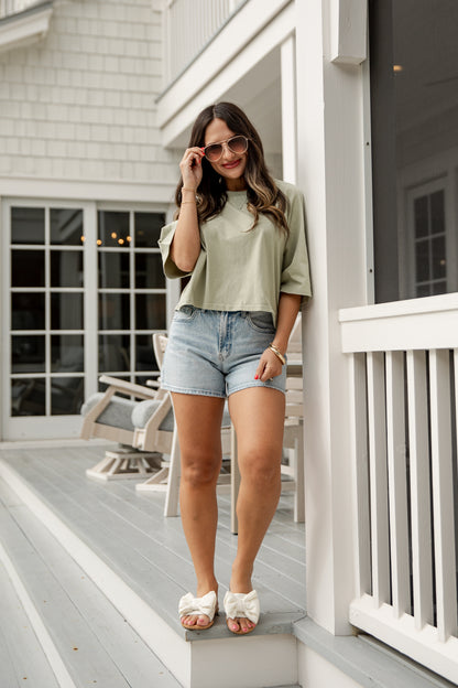 Woman standing on a porch wearing a green top and denim shorts.