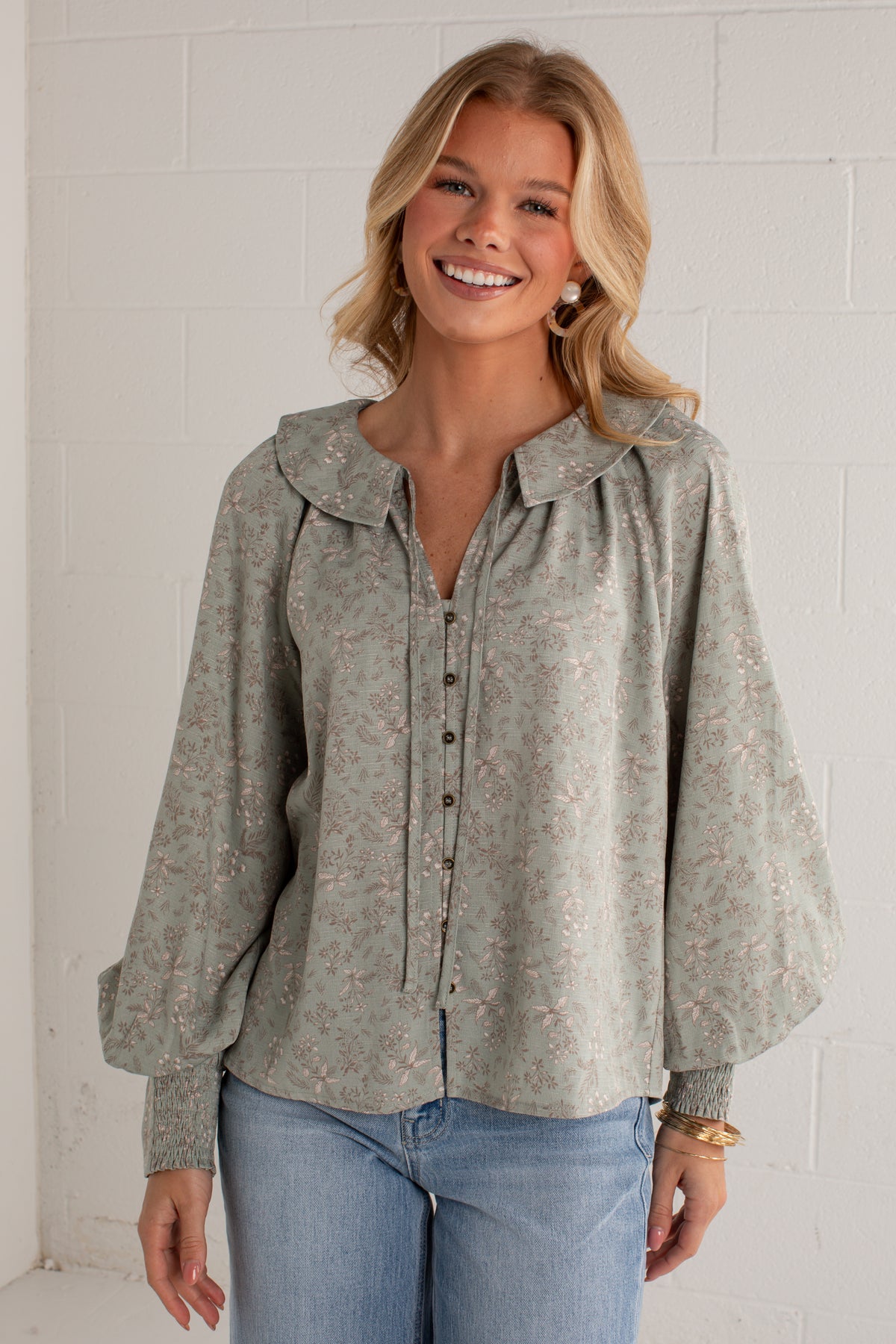 A woman wearing a sage green long-sleeve blouse with a delicate pink and white floral pattern, paired with light blue jeans, standing against a white brick wall.
