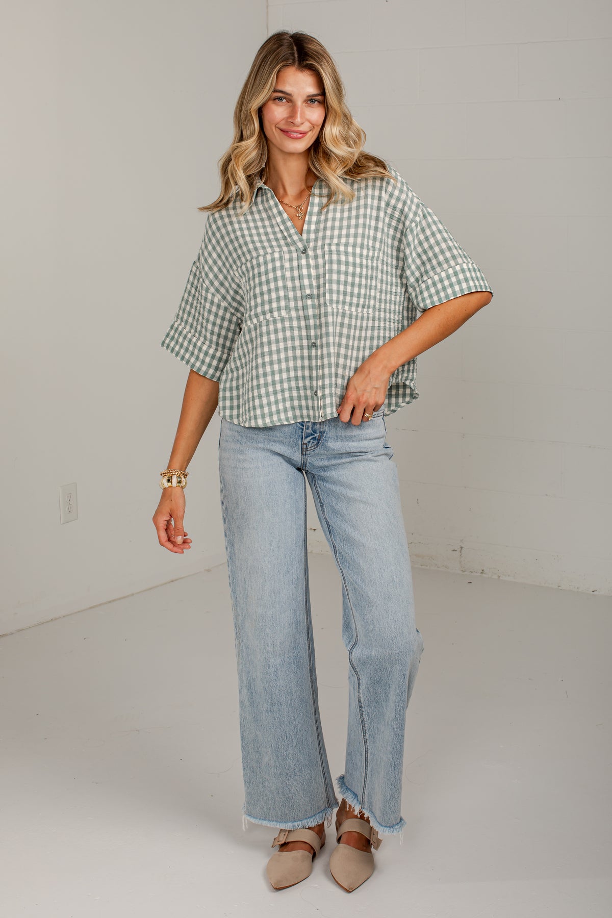 A woman modeling a light green and white gingham short-sleeve button-up shirt paired with light-wash wide-leg jeans and beige pointed-toe flats, standing against a plain off-white studio wall.
