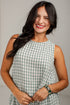 A smiling woman with long dark hair wearing a sleeveless green and white gingham dress, standing against a light gray background.