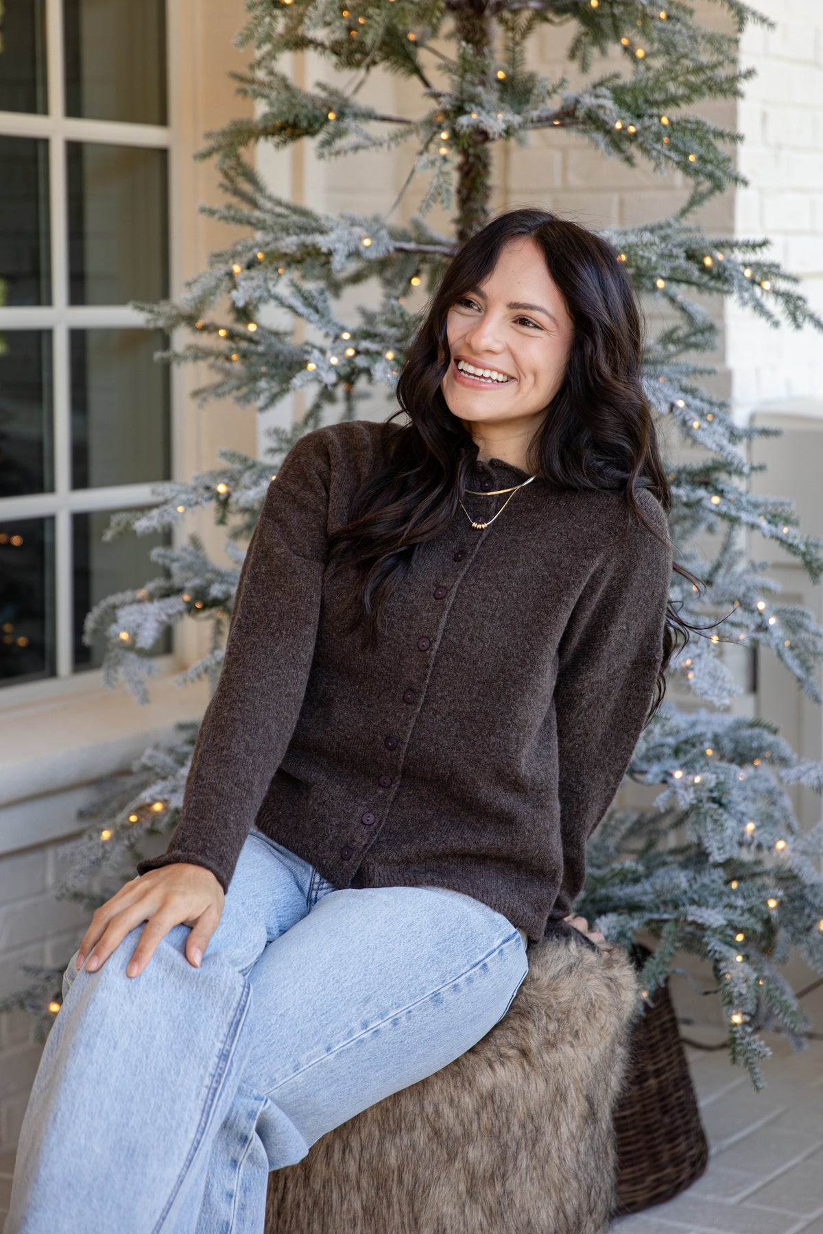 A woman with long dark hair sits smiling in front of a decorated Christmas tree, wearing a dark brown knit sweater and light blue jeans.