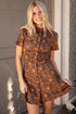A smiling blonde woman wearing a brown leopard-print short-sleeve button-up dress, standing outdoors in front of a beige garage door and brick wall.