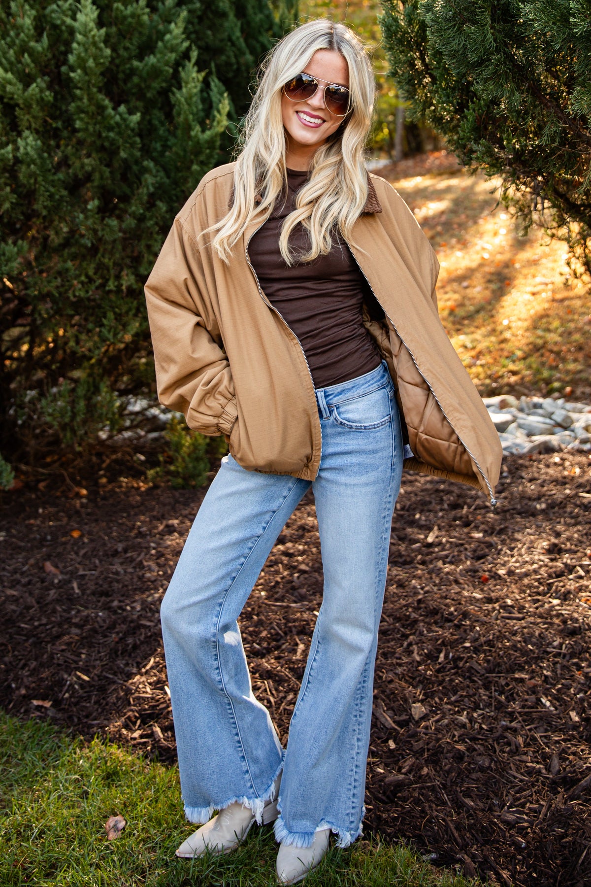 A woman posing outdoors in a tan oversized jacket, brown ribbed top, light blue flared jeans, and white sneakers, set against a background of green trees and mulch.