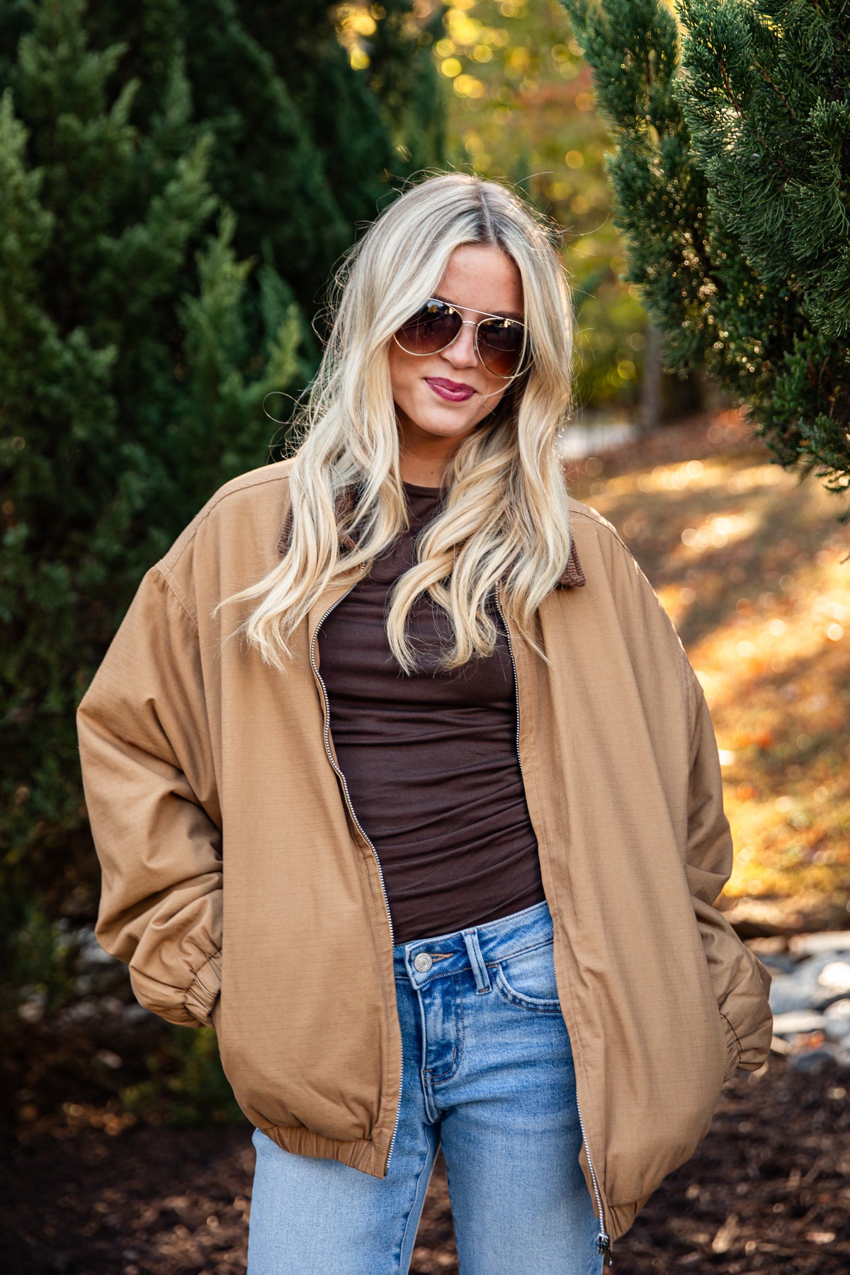 A woman wearing an oversized tan bomber jacket over a dark brown top and blue jeans, standing outdoors in front of green trees.