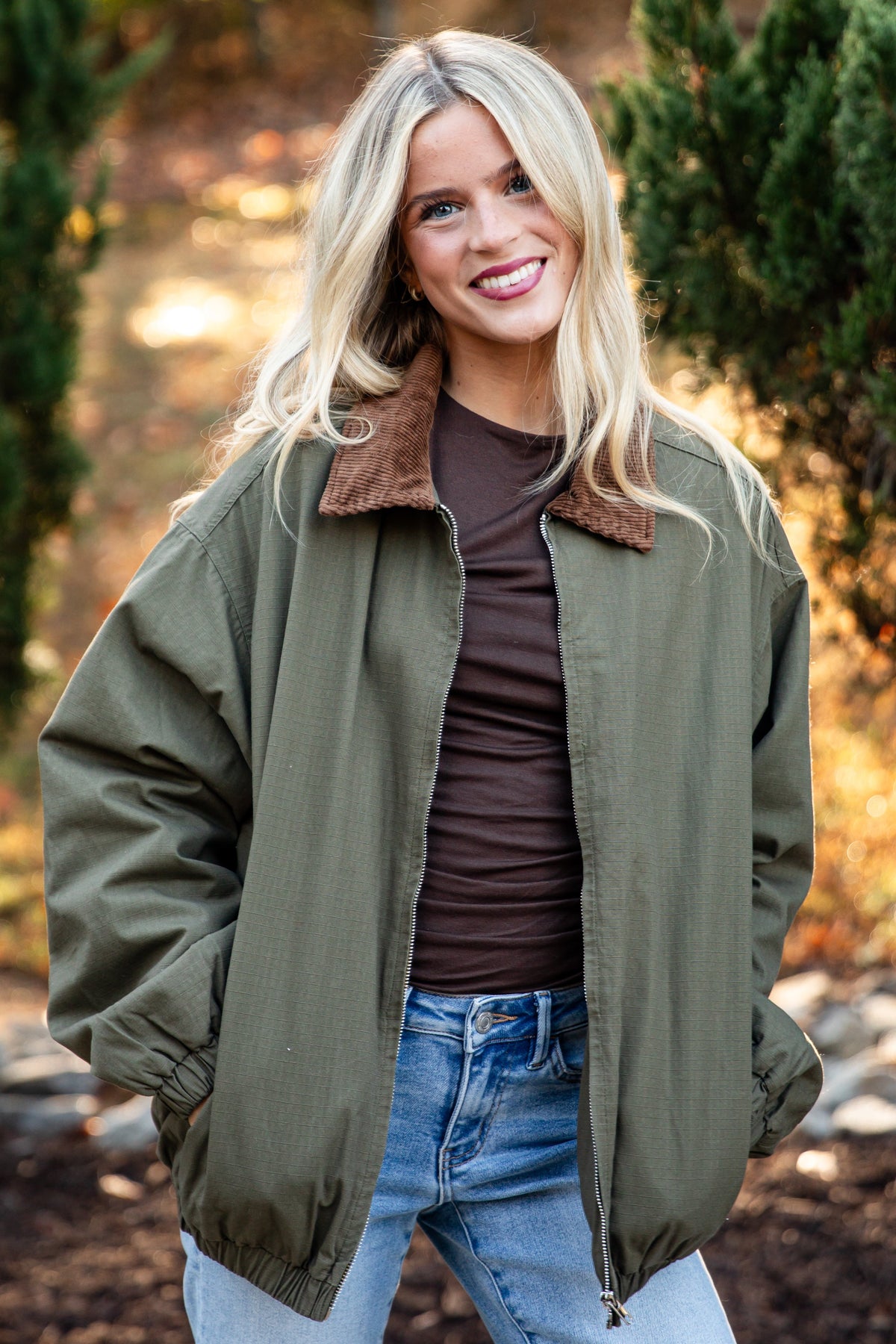 A woman standing outdoors in a wooded setting, wearing an olive green bomber-style jacket with a brown sherpa collar over a dark brown top and light blue jeans.