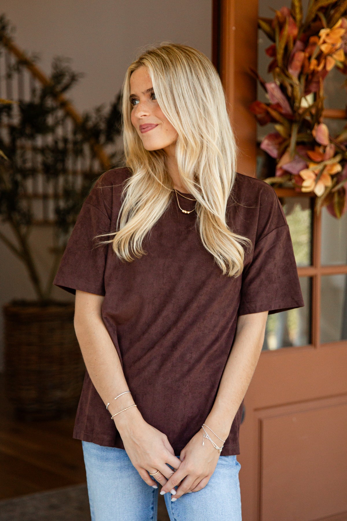 A blonde woman wearing a dark brown short-sleeve t-shirt and light blue jeans, standing in a home entryway with autumn decorations.
