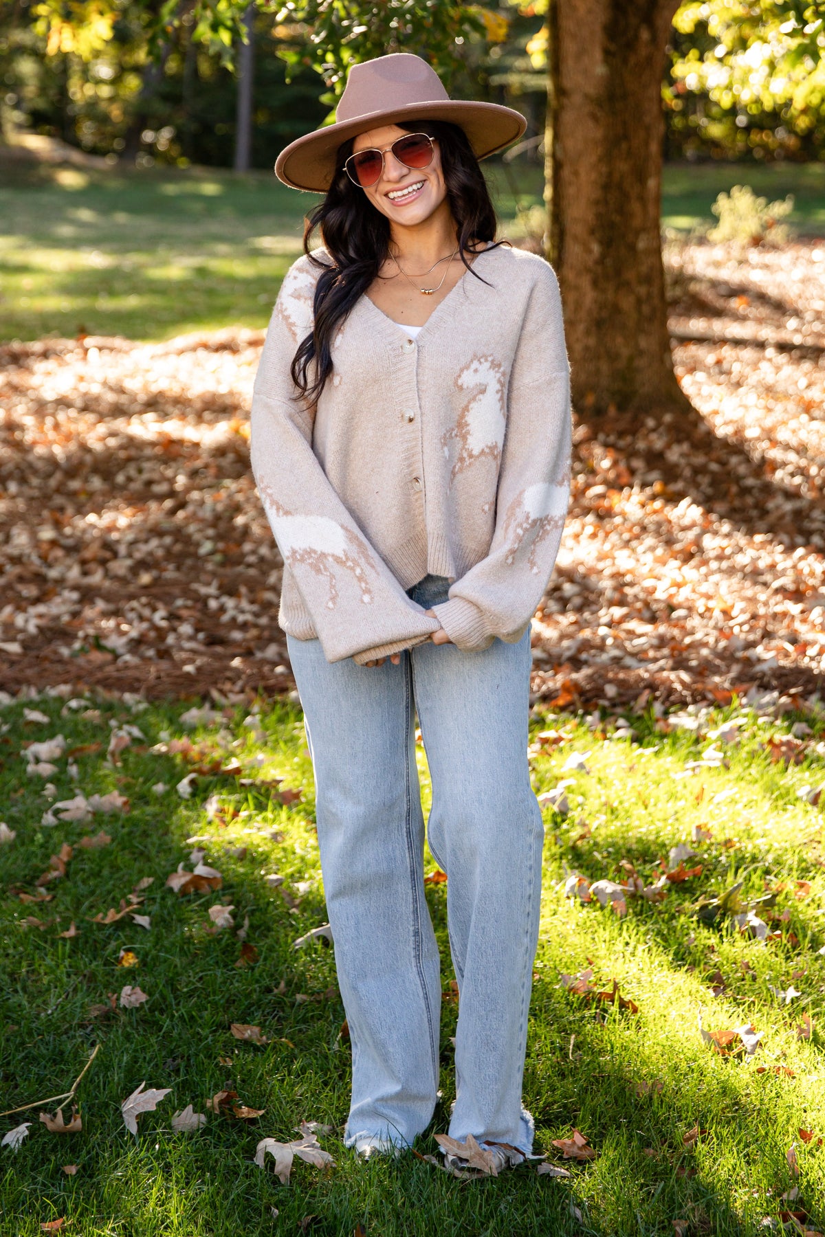 A woman stands outdoors in a park wearing a beige distressed cardigan, light blue jeans, a brown wide-brim hat, and sunglasses.