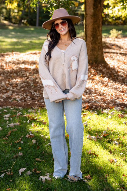 A woman stands outdoors in a park wearing a beige distressed cardigan, light blue jeans, a brown wide-brim hat, and sunglasses.