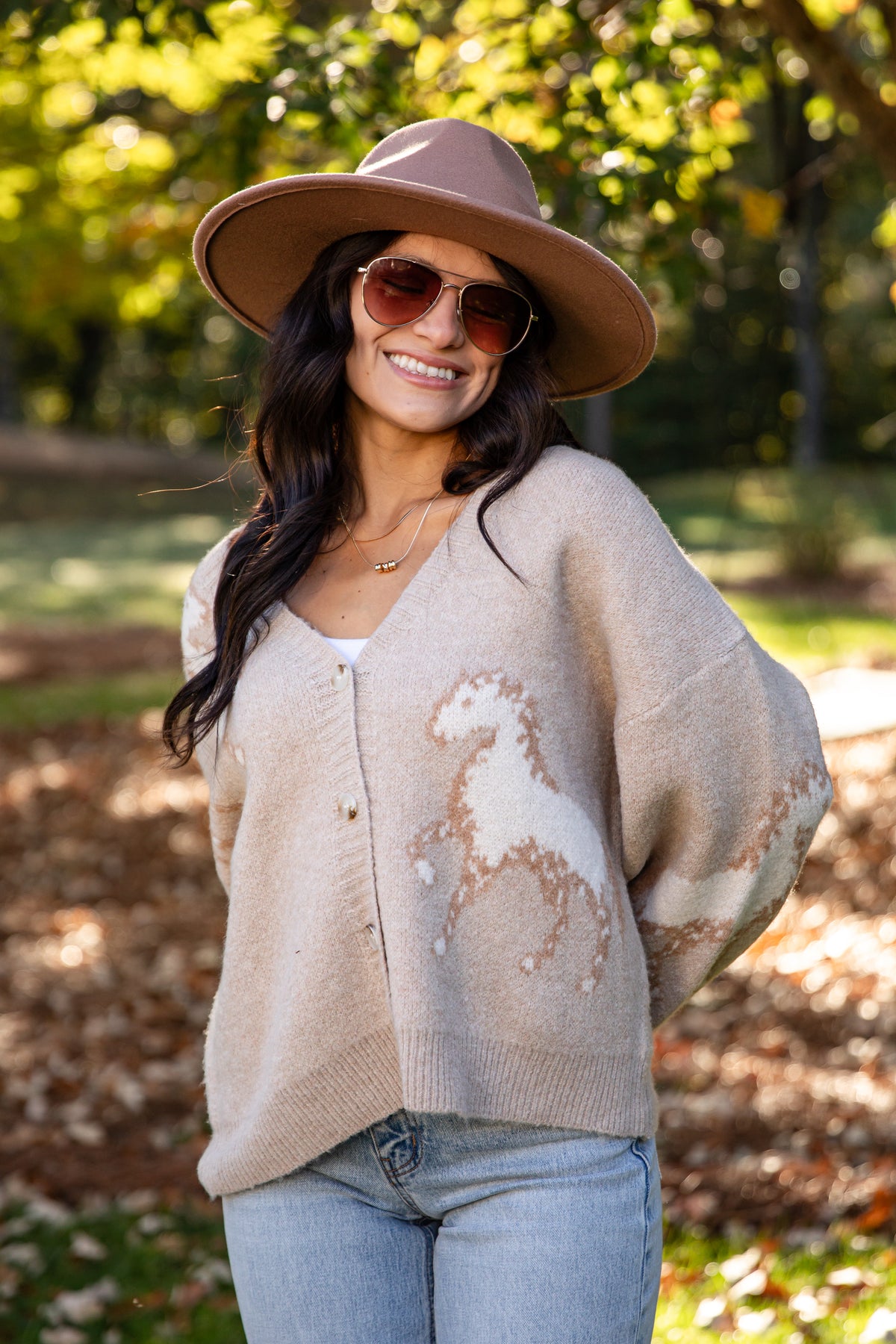 A woman wearing a beige knit cardigan with a white horse motif, a brown wide-brim hat, and sunglasses, standing outdoors in a park with trees and fallen leaves in the background.