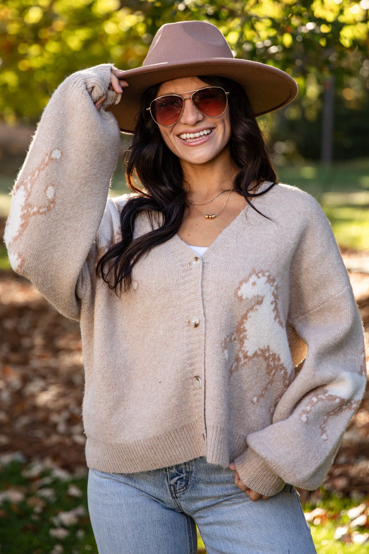 A woman wearing a beige distressed knit cardigan, a brown wide-brim hat, and sunglasses, posing outdoors in a park with autumn trees and fallen leaves.