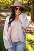 A woman outdoors wearing a beige knit cardigan with white distressed patches, a dusty pink wide-brim hat, and sunglasses, standing in a sunny park with trees and fallen leaves.