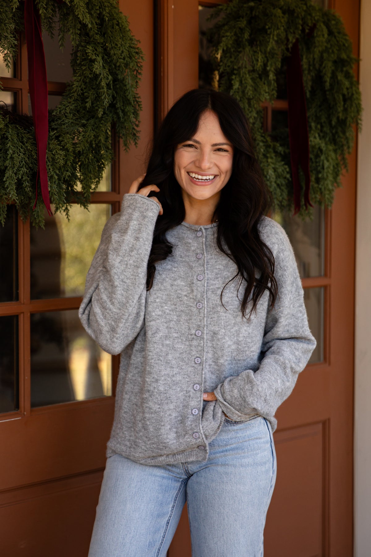 A woman smiling and posing in front of a rustic brown wooden door decorated with green holiday wreaths. She is wearing a light gray heathered button-up cardigan sweater and light wash blue jeans.