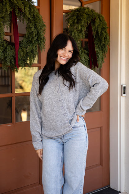 A woman wearing a light gray knit button-front cardigan and light blue jeans stands smiling in front of a brown wooden door decorated with green wreaths.