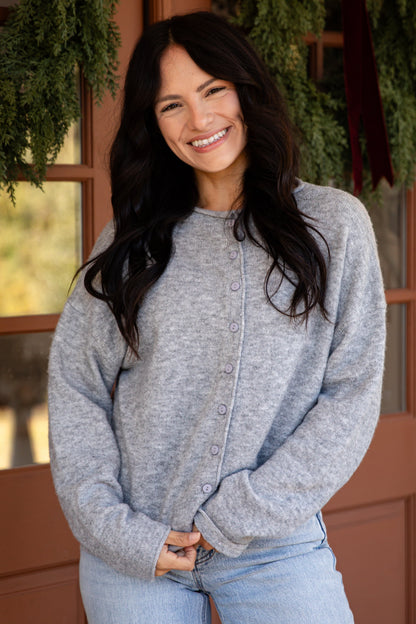 A smiling woman with long dark hair stands in front of a brown door decorated with green wreaths, wearing a light gray knit cardigan and light blue jeans.