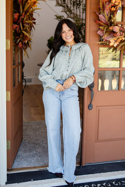 A woman stands in a doorway wearing a light sage green hooded button-up shirt and light blue wide-leg jeans. The setting features a rustic brown door with glass panes, fall wreaths, and a gray rug.