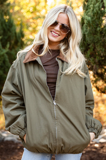 A woman wearing an olive green zip-up jacket with a brown sherpa collar, styled with a brown top and light blue jeans, standing outdoors in a wooded setting.