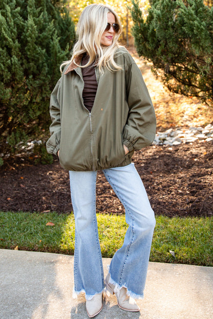 A woman stands outdoors on a concrete path wearing an olive green zip-up jacket over a dark striped shirt, light blue flared jeans, and white sneakers, with trees and sunlight in the background.