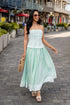 Woman in a light green dress walking on a street with buildings and plants in the background