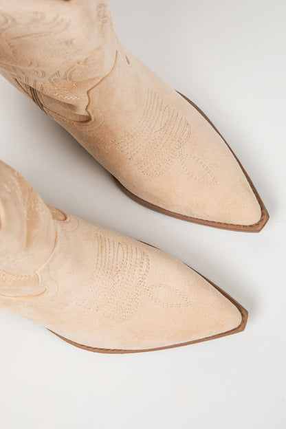 A pair of pointed-toe western boots in light beige suede with decorative embossed stitching, shown from a top-down angle on a light gray background.