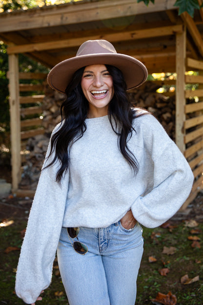 A woman smiling outdoors in front of a wooden structure, wearing a light beige wide-brim hat, a fuzzy light gray oversized sweater, and light blue jeans.