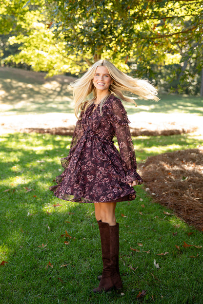 A woman outdoors on grass wearing a dark floral long-sleeve mini dress with a tie waist and brown knee-high boots. Trees and sunlight are visible in the background.