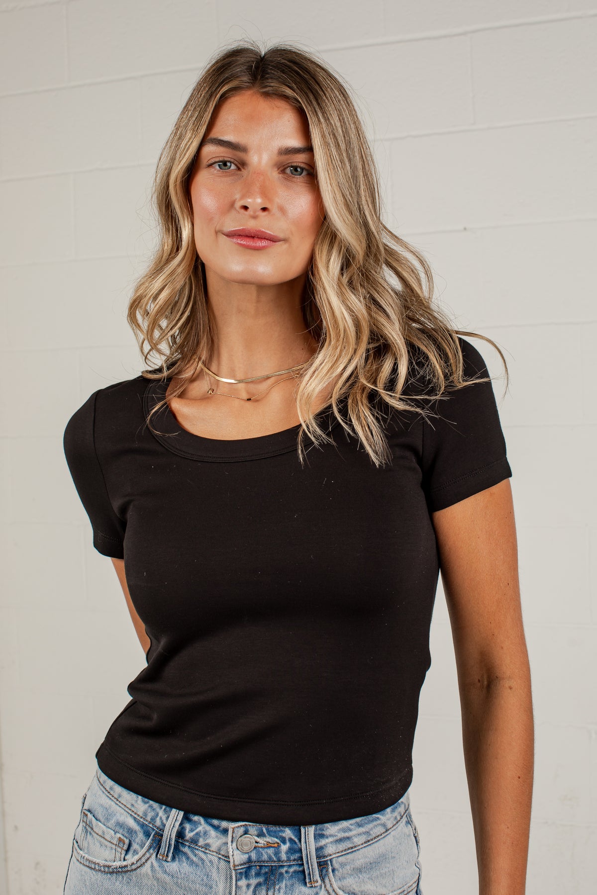 A woman with blonde-highlighted hair wearing a fitted black short-sleeve scoop-neck t-shirt and light-wash blue jeans, standing against a white brick wall.