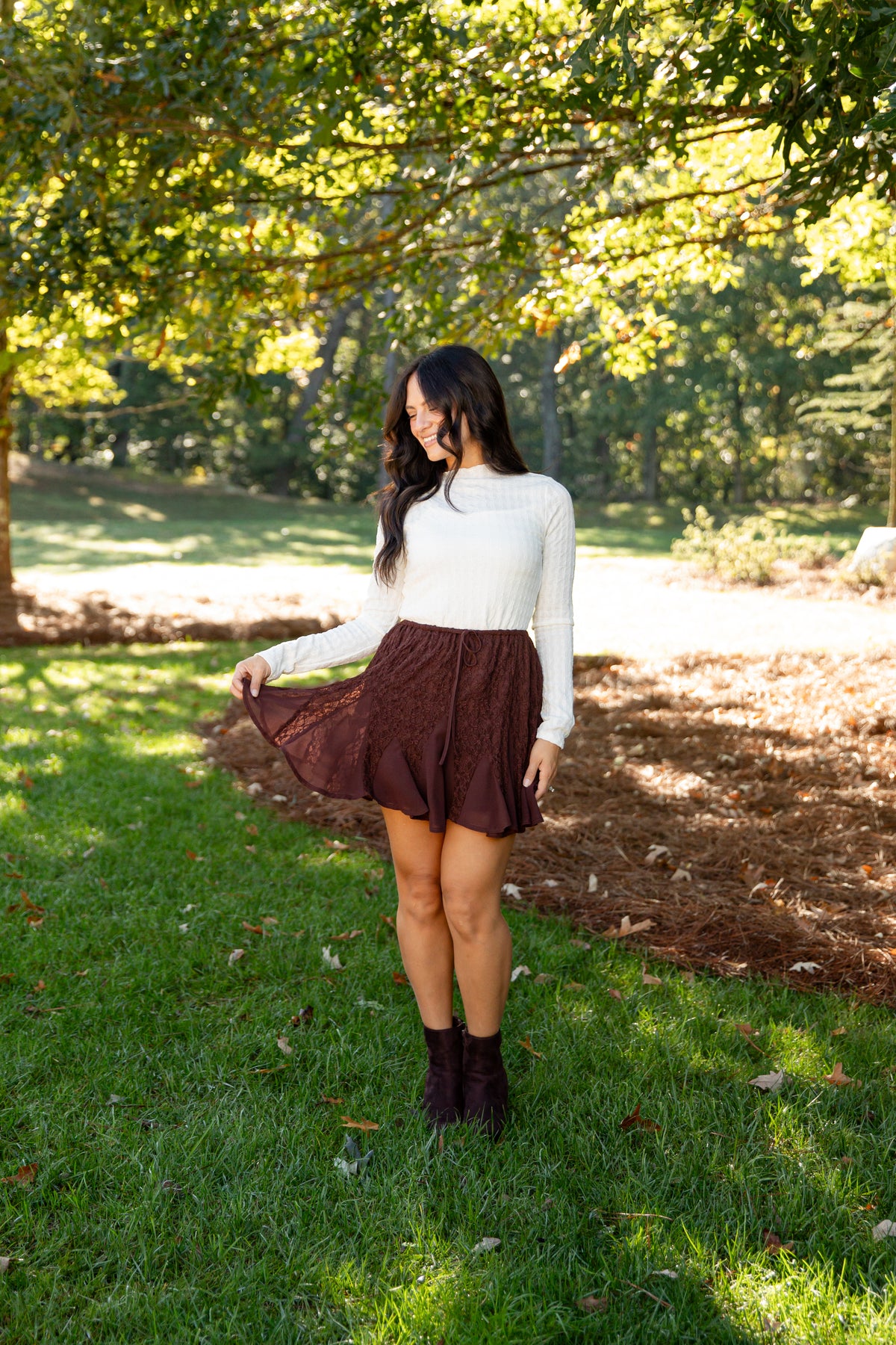 A woman stands on a grassy lawn under a tree, wearing a white ribbed long-sleeve top, a brown pleated mini skirt, and dark brown ankle boots.
