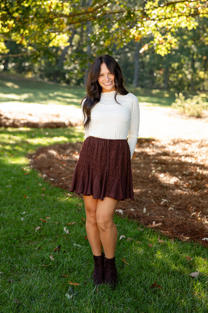 A woman standing outdoors in a grassy park area under trees, wearing a white ribbed turtleneck sweater and a dark brown pleated skirt, paired with dark brown ankle boots.