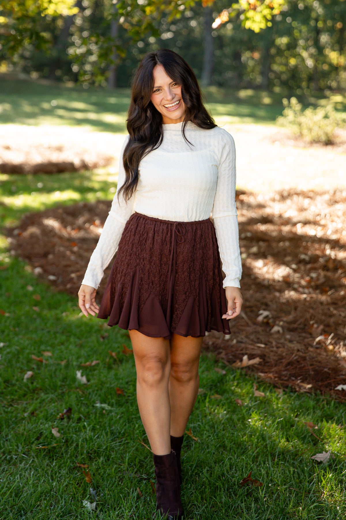 A woman standing outdoors on a grassy lawn wearing a cream ribbed long-sleeve mock neck top, a dark brown textured pleated mini skirt, and dark brown ankle boots.