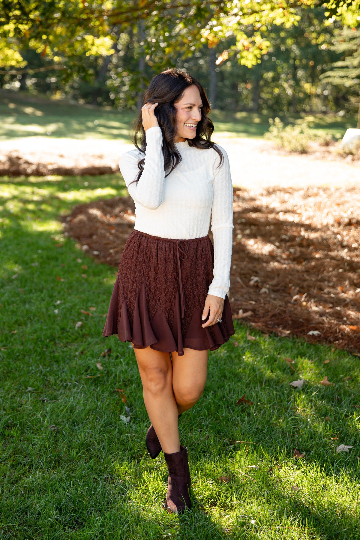 A woman stands outdoors on a grassy park path wearing a white long-sleeve knit top and a dark brown textured pleated mini skirt with dark brown ankle boots.