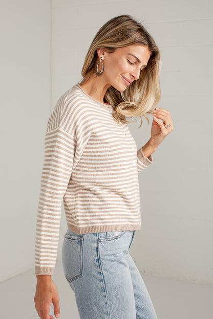 A woman wearing a beige and white horizontally striped knit sweater and light blue jeans, standing in a bright, minimalist room with white paneled walls.