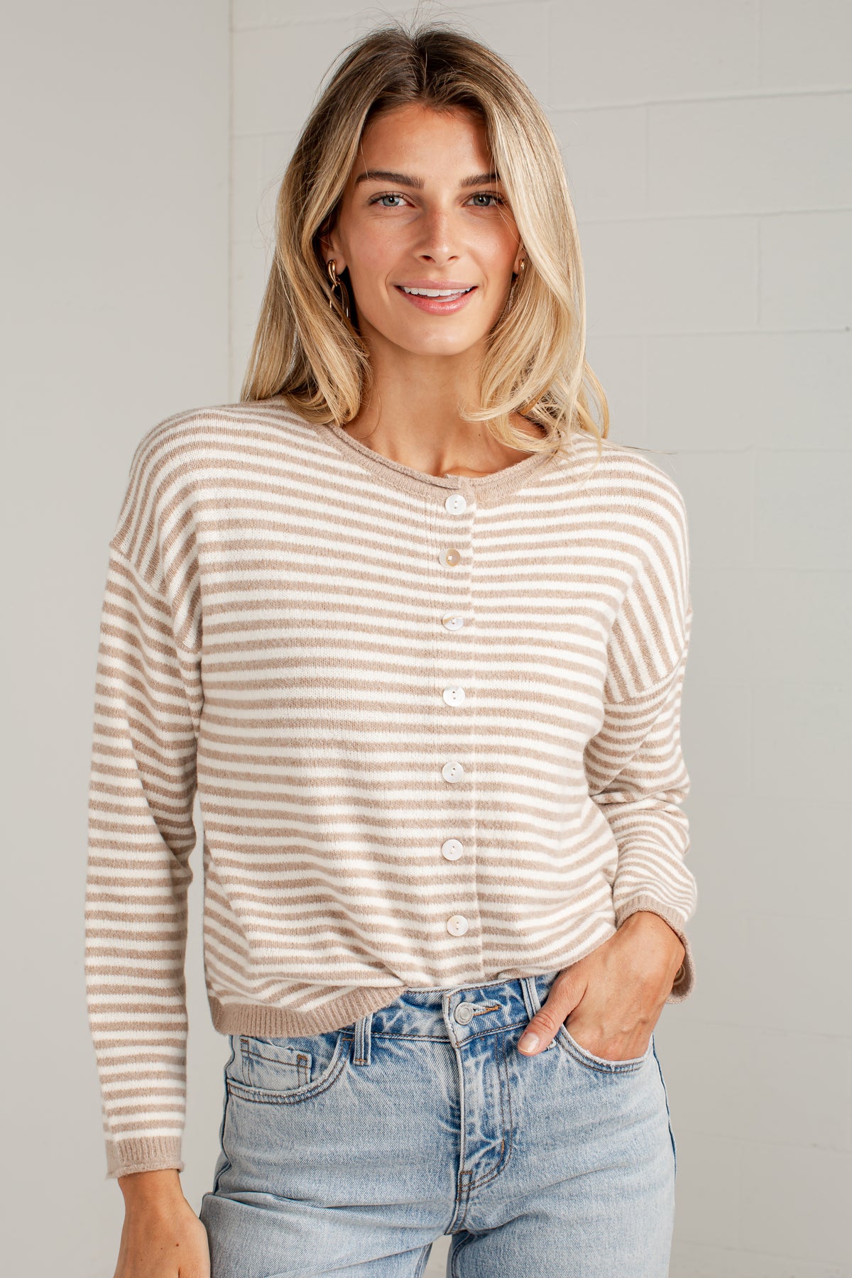 A woman wearing a beige and white horizontally striped button-up cardigan sweater with light blue jeans, standing against a light gray brick wall.