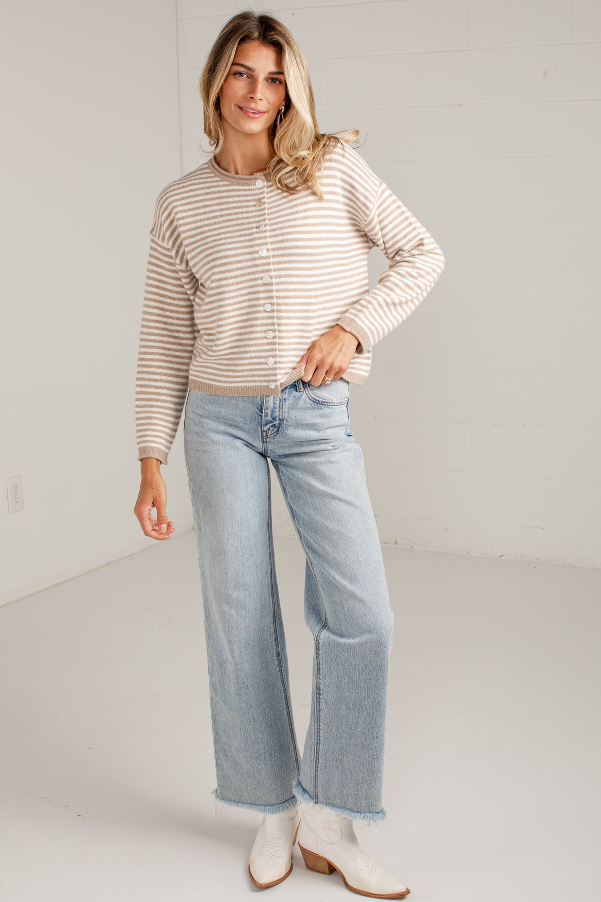 A woman modeling a beige and white striped long-sleeve button-up cardigan with light-wash wide-leg jeans and white ankle boots, standing against a white studio wall.