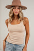 A woman wearing a beige scoop-neck ribbed tank top, blue jeans, and a brown wide-brim fedora hat, posing against a white brick wall.