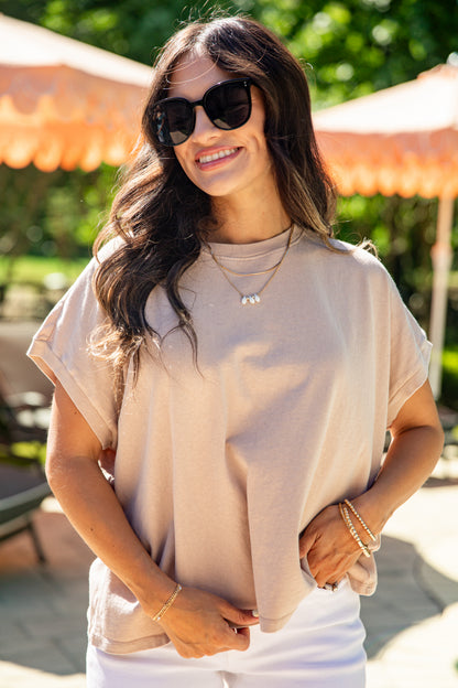 Woman wearing sunglasses and a beige top outdoors with greenery in the background