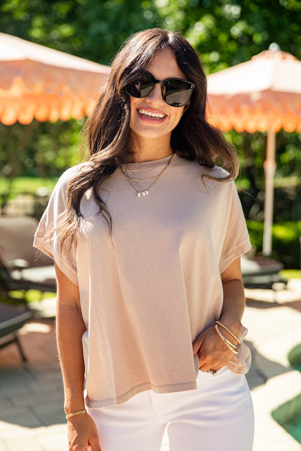Woman wearing sunglasses and a beige top in an outdoor setting with orange umbrellas.