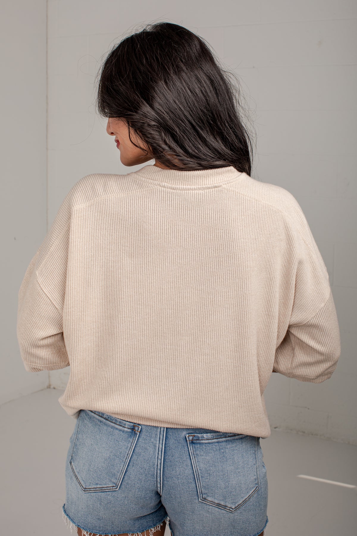 A woman shown from behind wearing a light beige ribbed knit long-sleeve top and blue denim jeans in a minimalist studio setting.