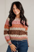 A smiling woman with long dark hair wears a striped knit sweater with horizontal bands of pink, orange, brown, and cream, paired with blue jeans against a white brick wall.