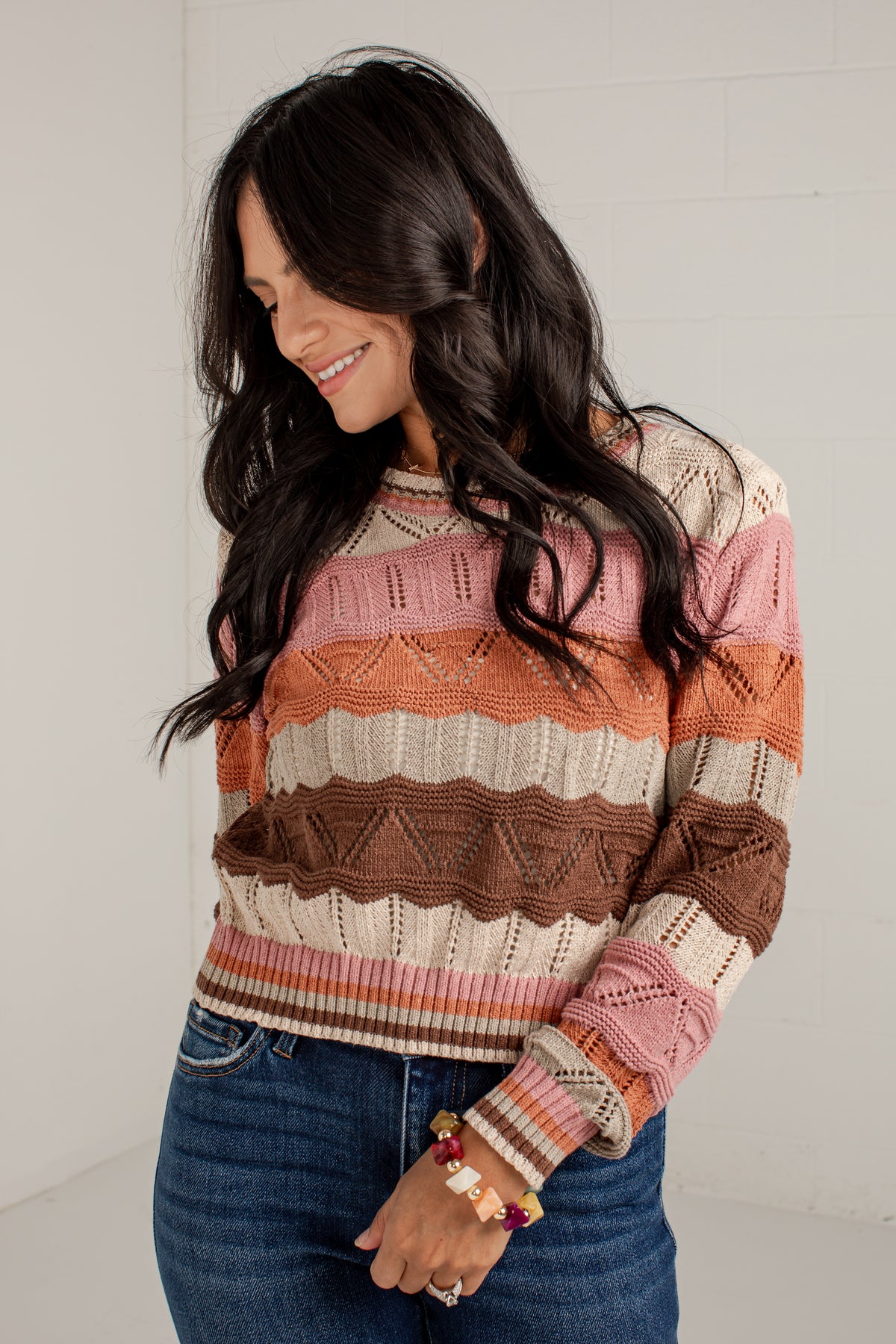 A woman wearing a long-sleeve knit sweater with horizontal stripes in pink, orange, brown, and cream, paired with blue jeans.