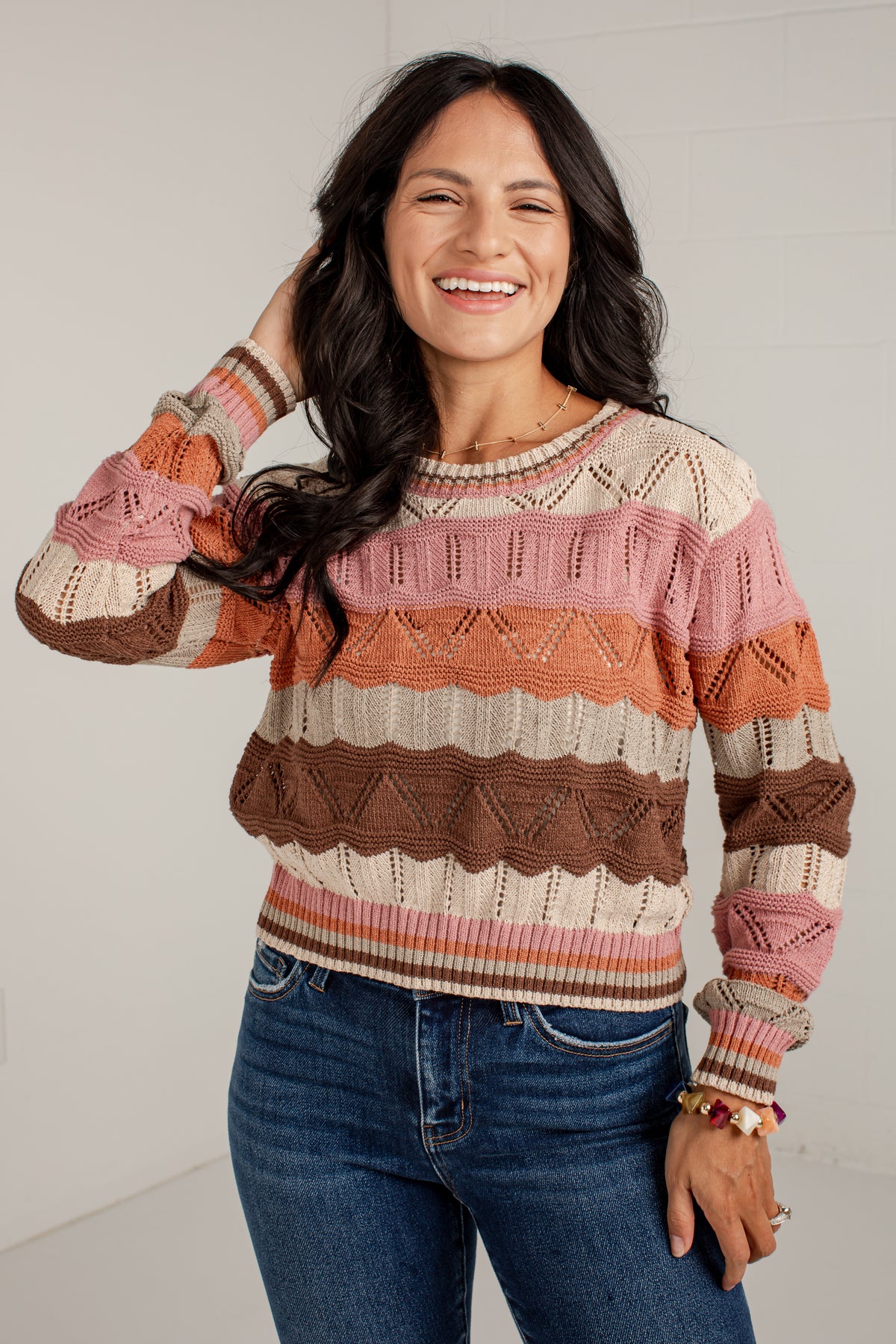 A woman smiling and wearing a long-sleeve knit sweater with horizontal stripes in pink, brown, orange, and cream, paired with blue jeans against a light gray wall.