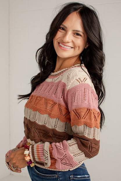A smiling woman with long dark hair wearing a multi-colored knit sweater with horizontal stripes in pink, orange, brown, and cream, paired with blue jeans.