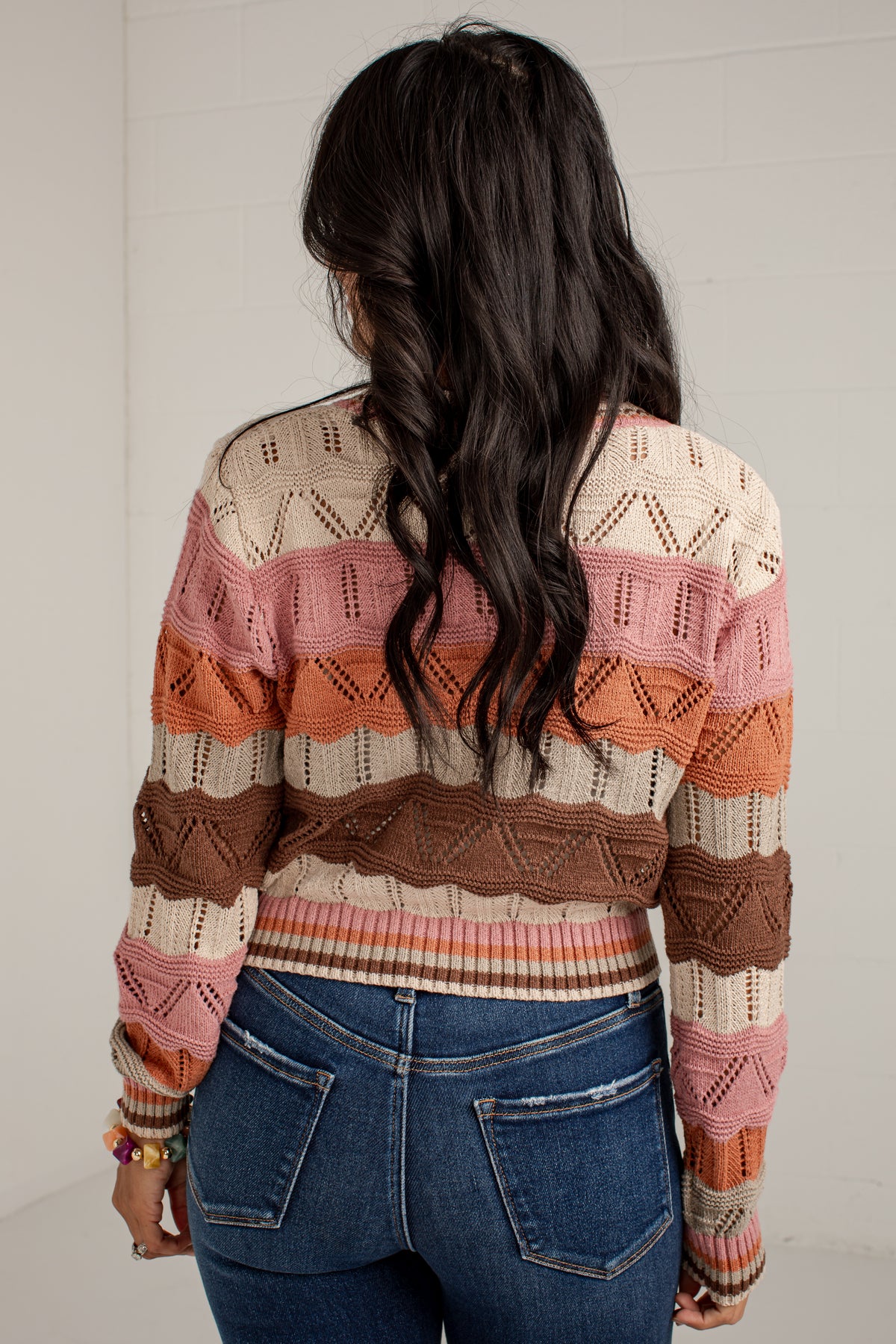 A woman viewed from behind wearing a striped knit cardigan with textured horizontal bands in cream, pink, orange, and brown, paired with blue jeans against a white brick wall.