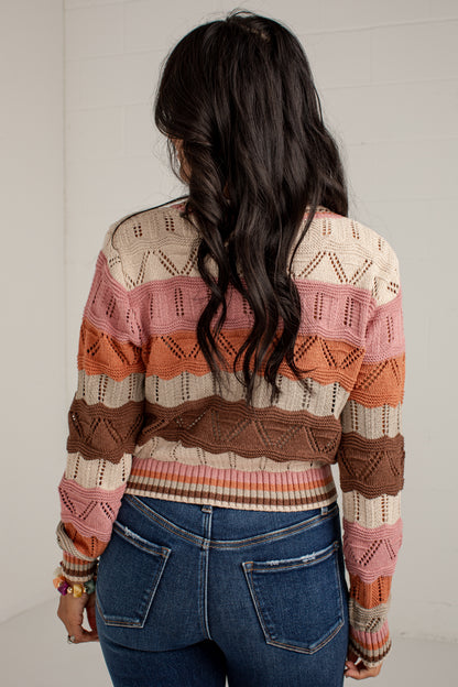 A woman viewed from behind wearing a striped knit cardigan with textured horizontal bands in cream, pink, orange, and brown, paired with blue jeans against a white brick wall.