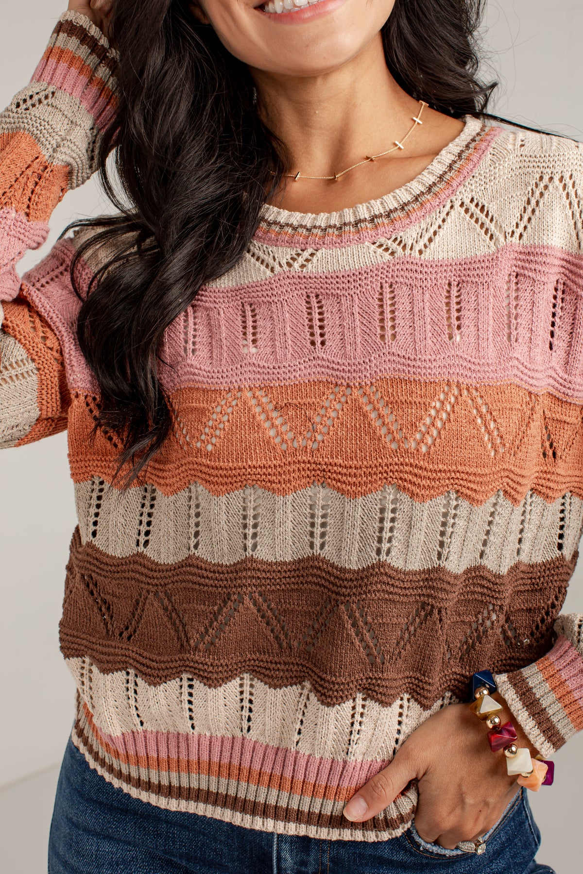 A close-up of a woman wearing a knit crewneck sweater with horizontal stripes in cream, dusty pink, terracotta orange, and brown, paired with blue jeans.