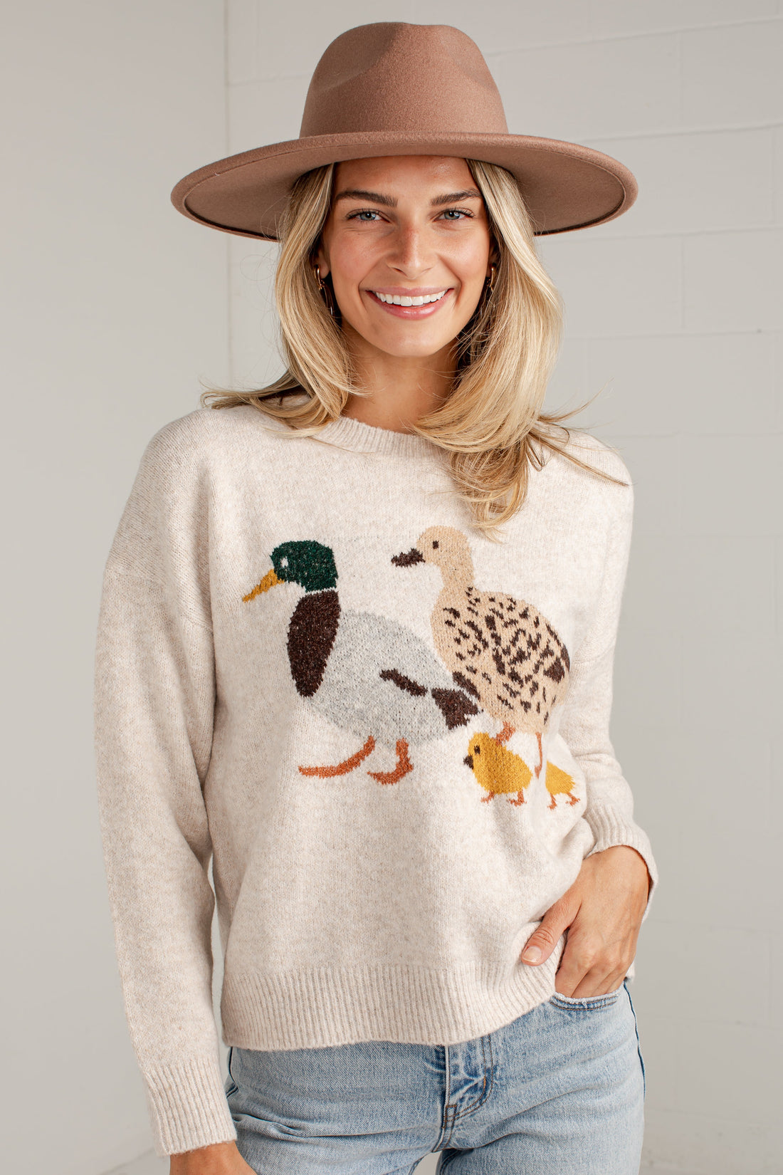 A woman wearing a beige knit sweater with a large embroidered duck family motif, paired with light blue jeans and a brown wide-brim hat, standing against a light gray wall.