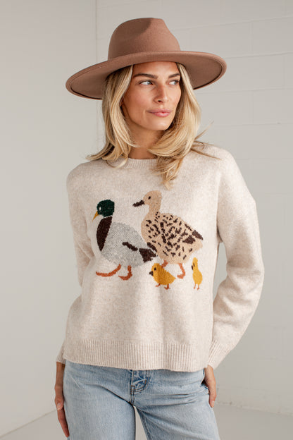 A woman wearing a cream-colored knit sweater with a large embroidered duck family motif on the front, paired with light blue jeans and a brown wide-brim hat, standing against a white brick wall.